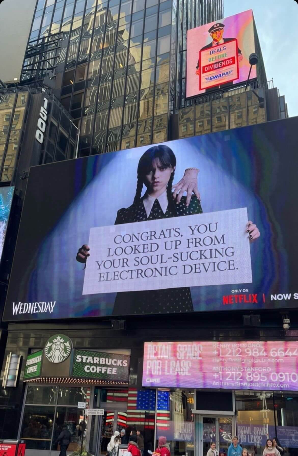 Outdoor banner promoting the Netflix series Wednesday, showing a photo of the main character holding a piece of paper which reads "Congrats, you looked up from your sould-sucking electronic device."