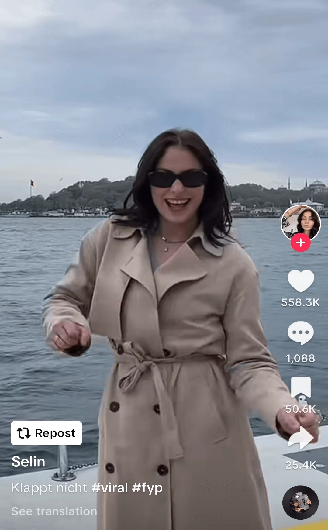Joyful woman in a stylish trench coat and sunglasses, smiling and enjoying a boat ride, shared in a social media post.