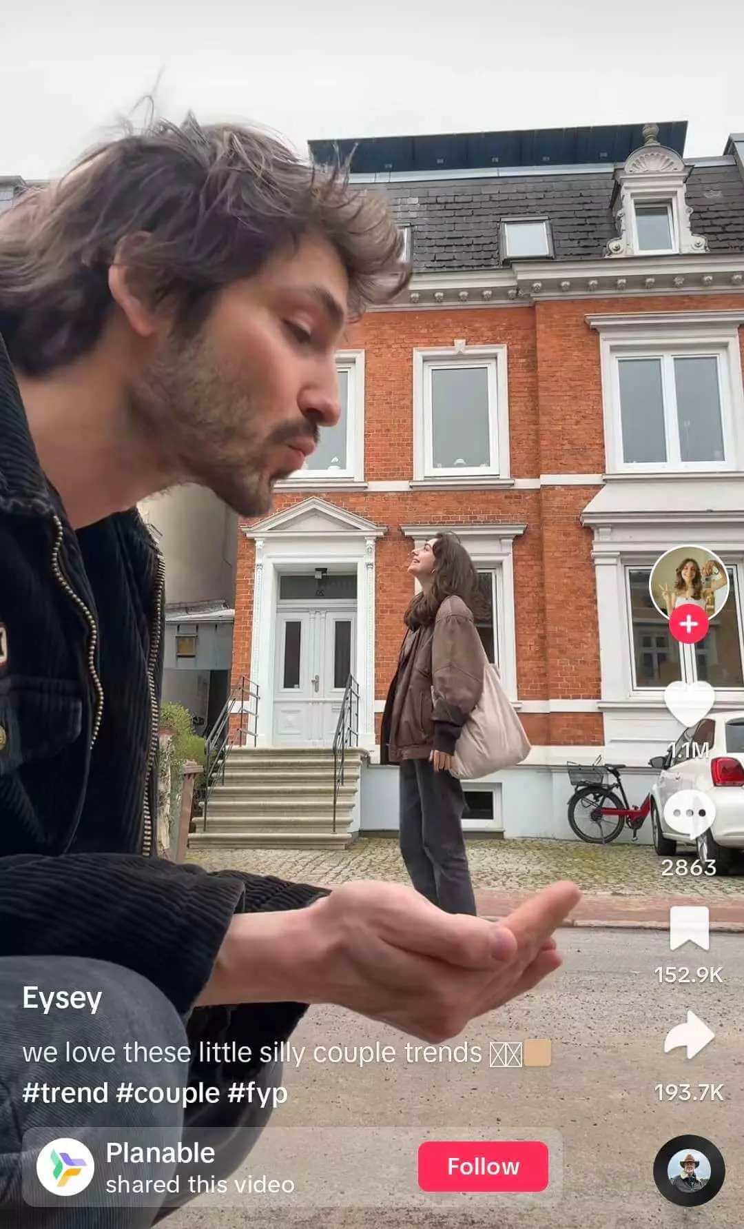 Man in foreground making a playful expression with a woman standing in the background on a residential street, featured in a light-hearted social media video about couple dynamics.