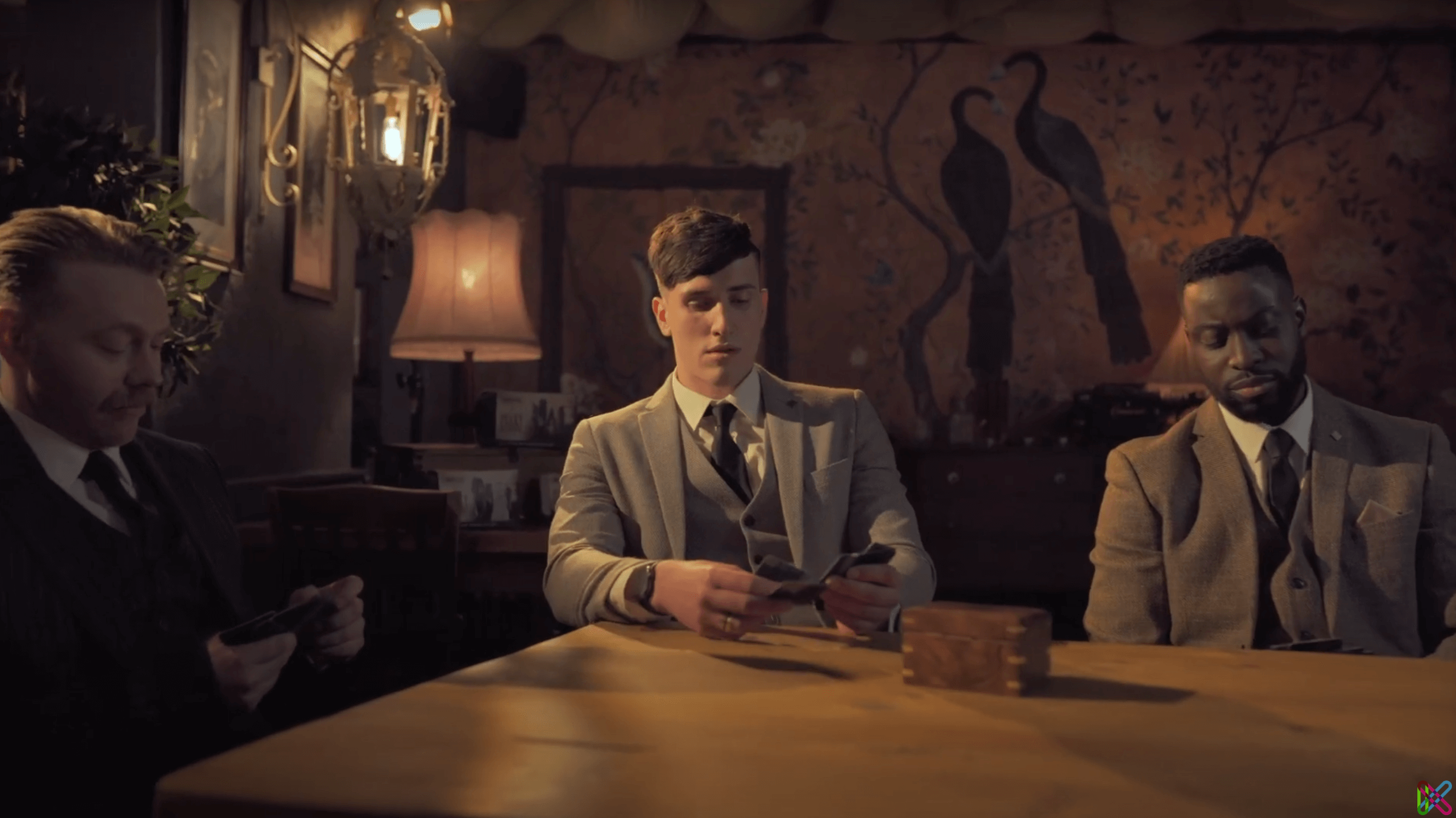 Three men in vintage suits sit at a table in a dimly lit room with a painted wall mural and vintage decor, in the style of the TV series Peaky Blinders.