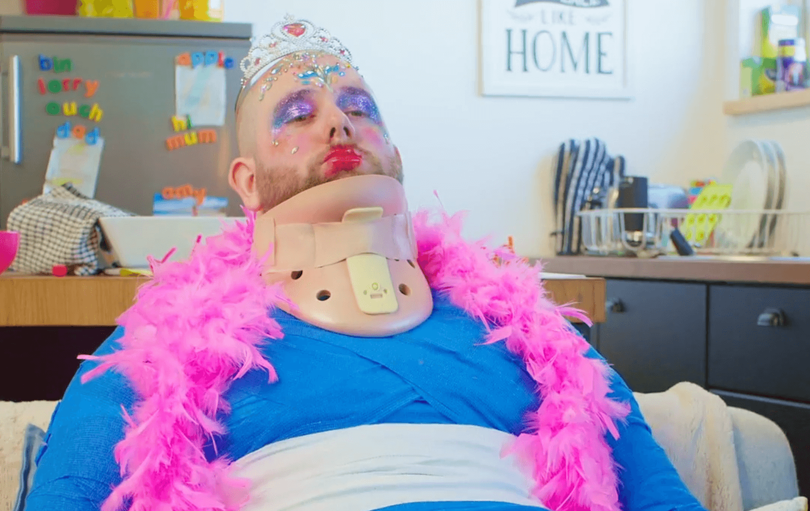 Man wearing a neck brace, pink feather boa, tiara, and heavy makeup, sitting in a kitchen with a sign "HOME" in the background.