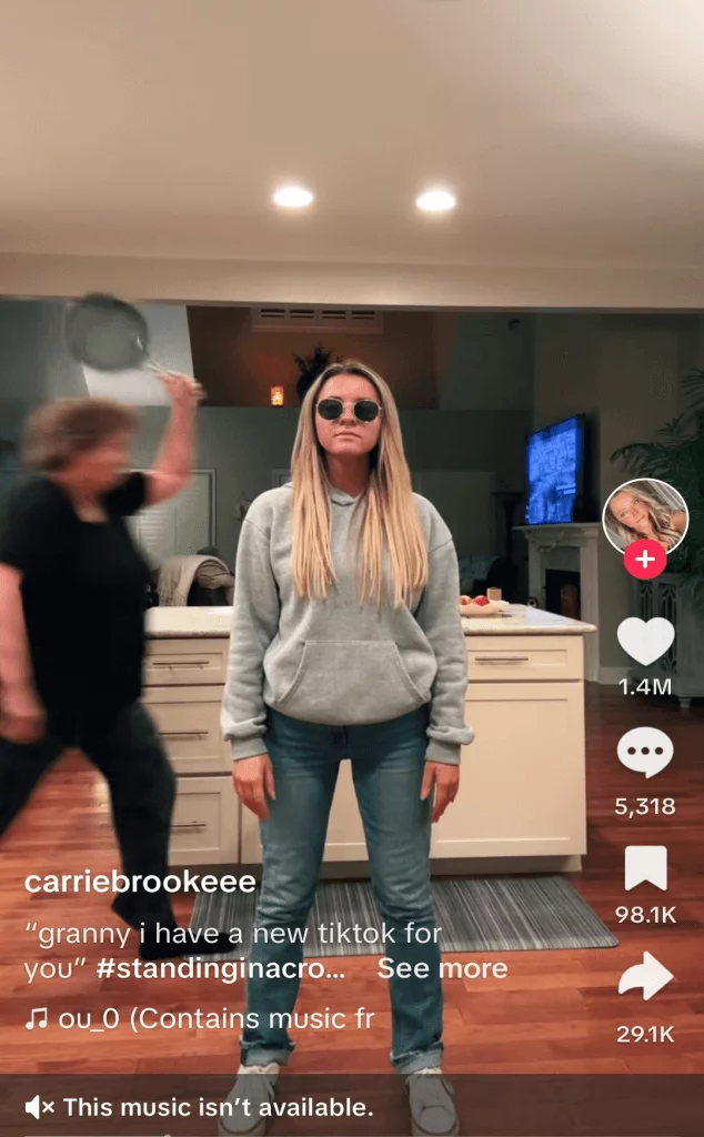 TikTok post of a woman standing in her kitchen, her grandmother is walking behind her with a frying pan.