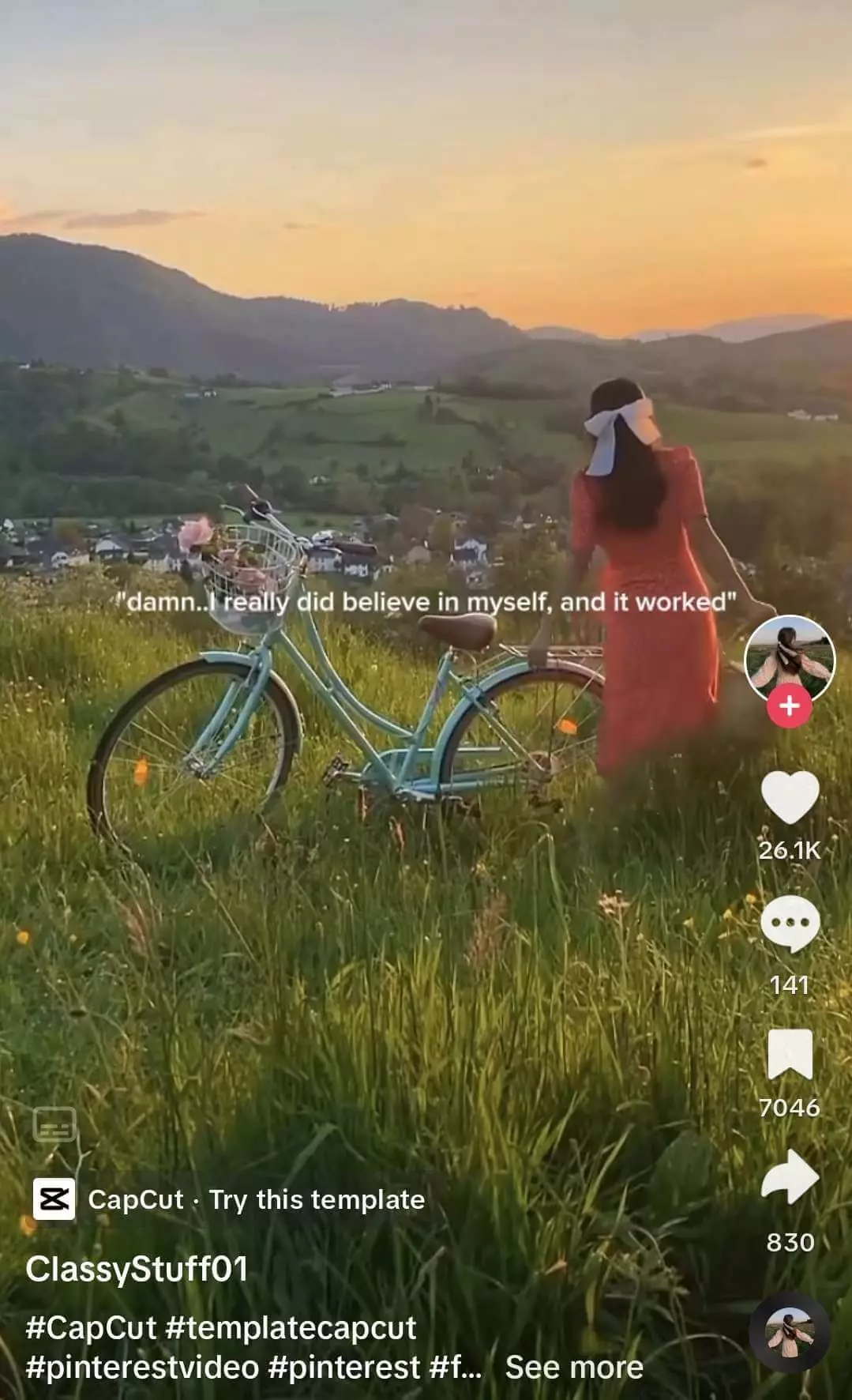 Woman in a red dress and white headscarf stands facing a sunset landscape, next to a vintage blue bicycle in a grassy field.