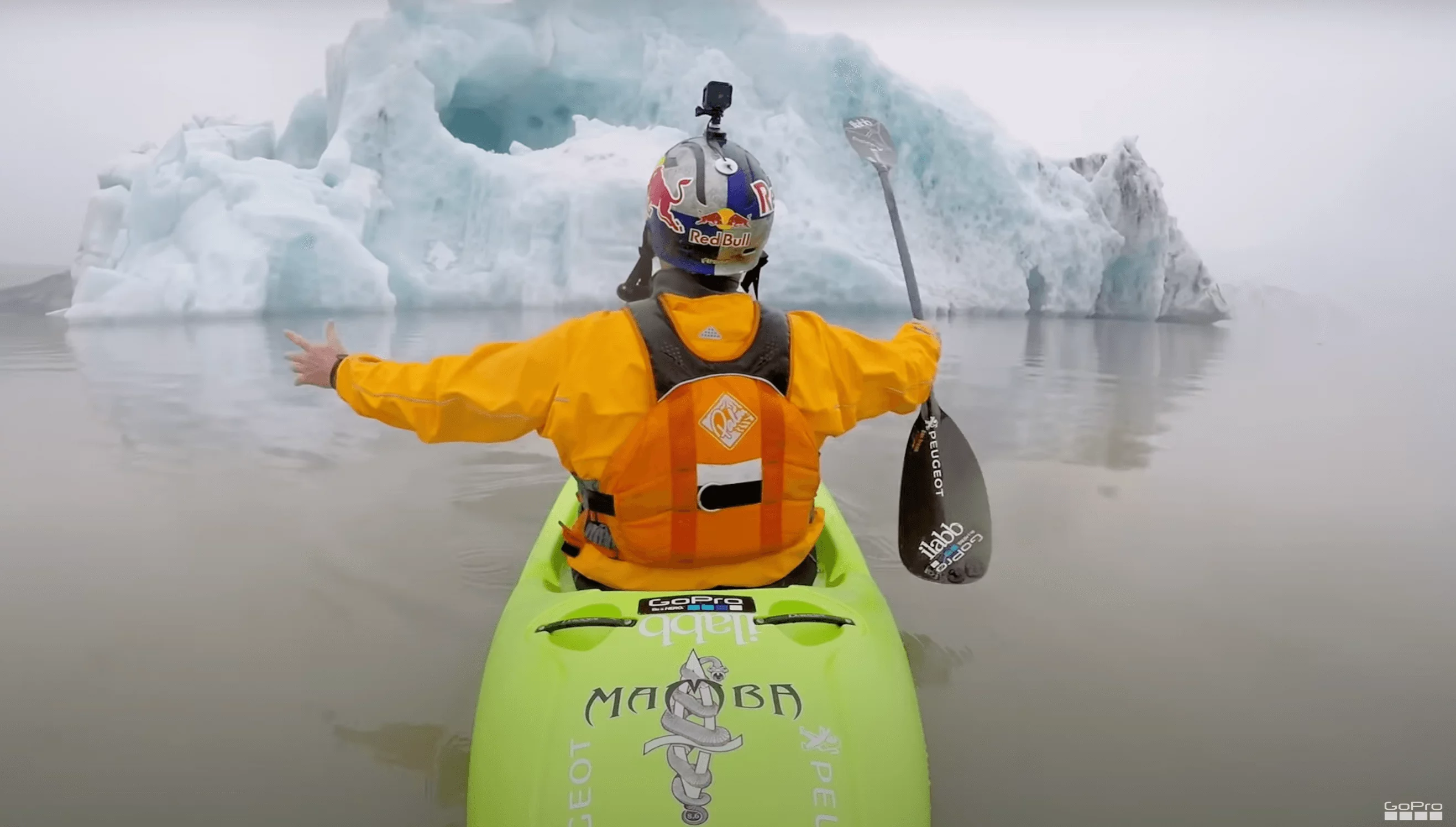 Kayaker in a bright orange jacket paddling towards a massive iceberg in foggy conditions, capturing the moment with a mounted GoPro.