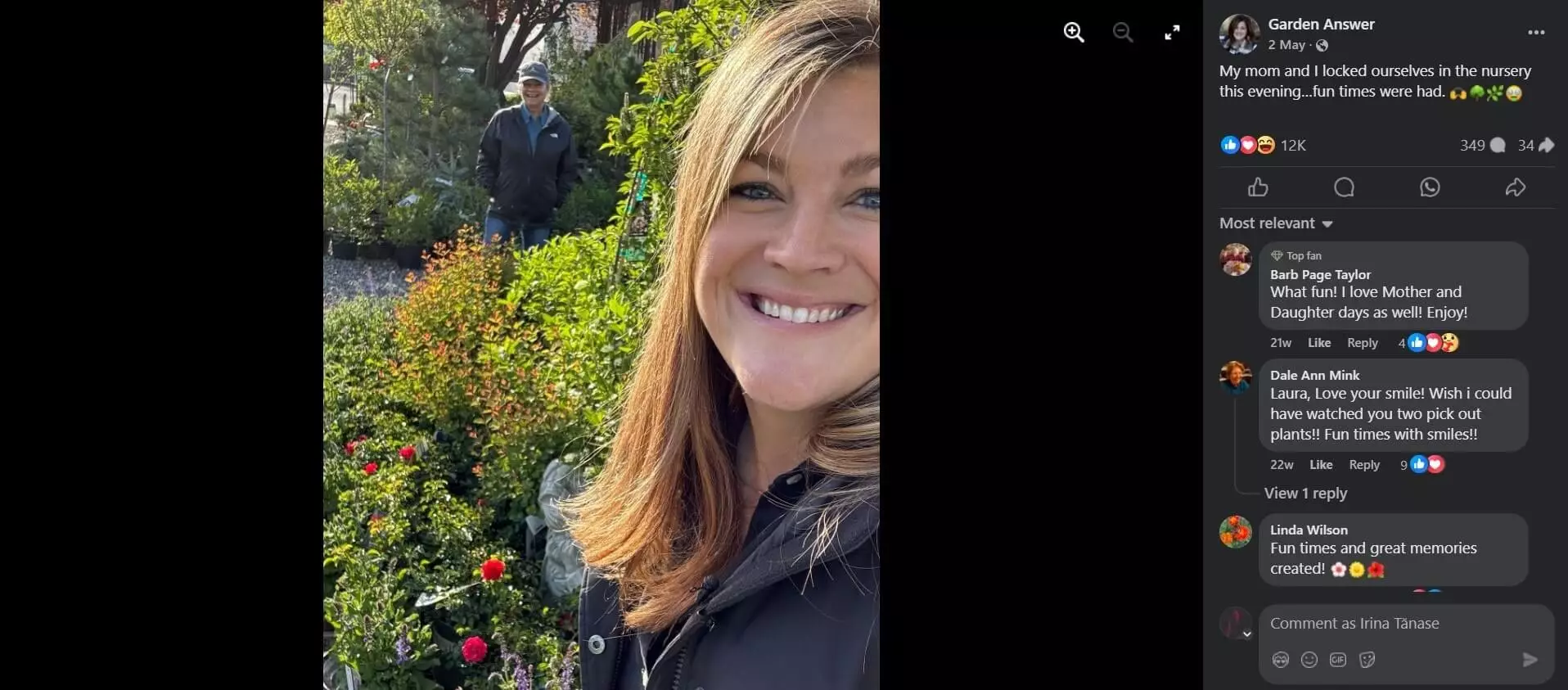 Selfie of a smiling woman at a plant nursery, surrounded by greenery and flowers, with another person in the background.